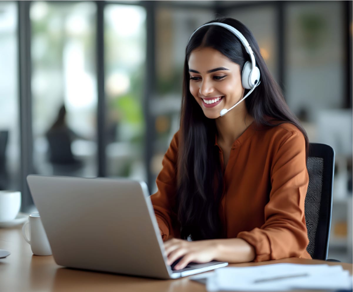 A woman wearing a headset sits at a desk, smiling and working on a laptop