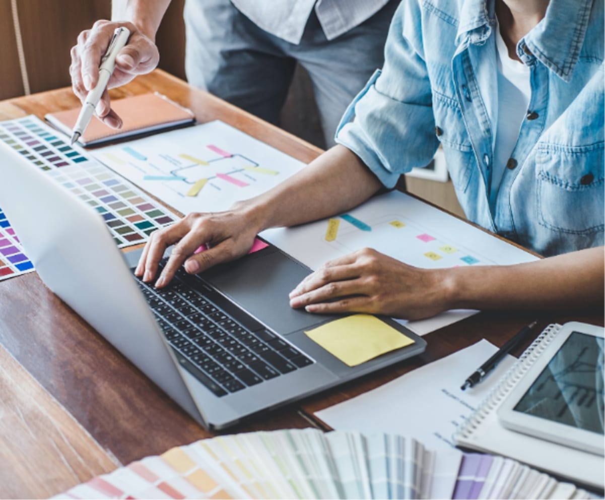 Two people work at a desk with a laptop, color swatches, and charts, discussing and pointing at design materials while planning a website project.
