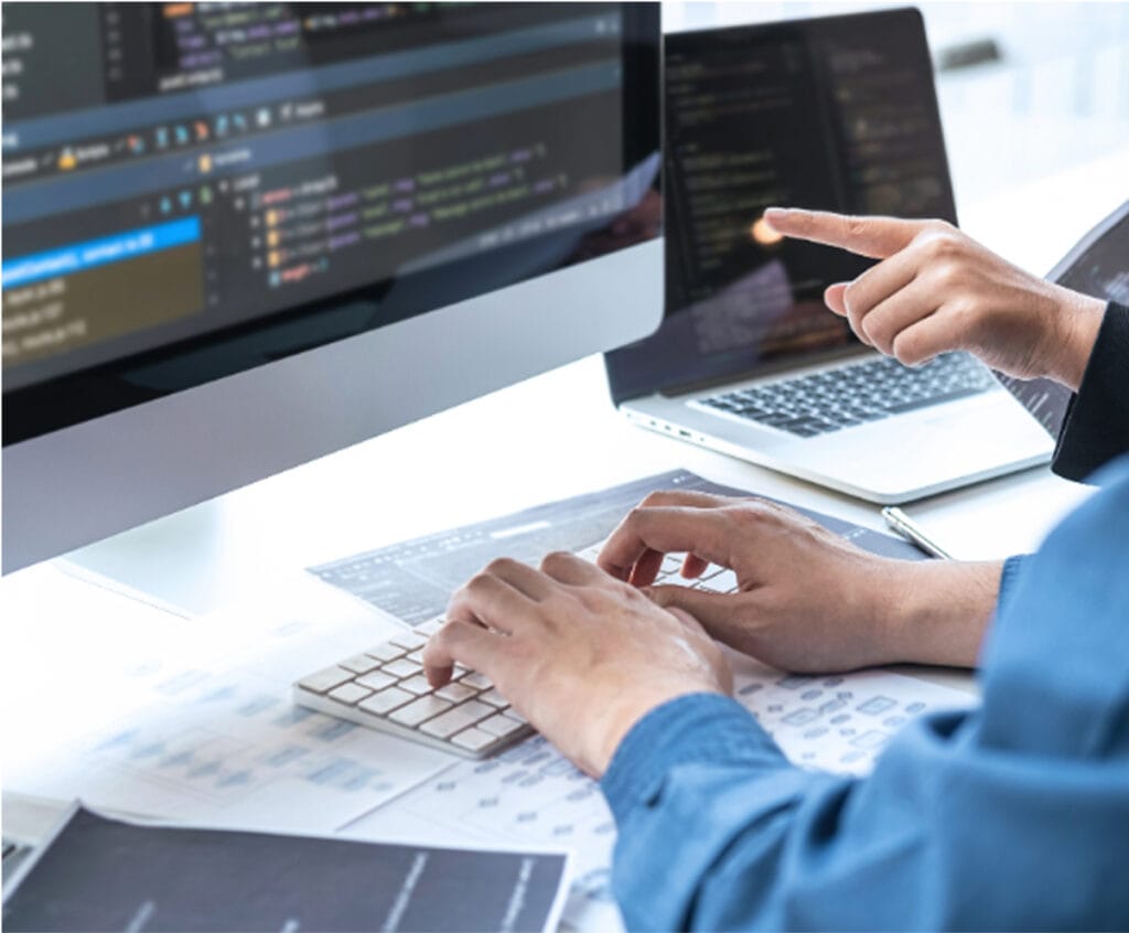 Two people work at a desk with computers and laptops displaying code
