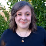 Sarah Gzemski has shoulder-length brown hair and a black top stands outdoors in front of green foliage, smiling at the camera.