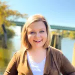 Ashley McLeroy has short blonde hair, smiling outdoors in front of a river, with a bridge and trees in the background.